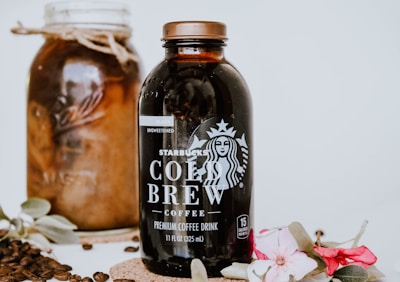 A bottle of Starbucks Cold Brew coffee is prominently displayed alongside a jar filled with coffee beans. Decorative flowers and leaves adorn the scene, adding elegance. Coffee beans are scattered around for an aesthetic touch.