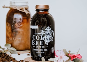 A bottle of Starbucks Cold Brew coffee is prominently displayed alongside a jar filled with coffee beans. Decorative flowers and leaves adorn the scene, adding elegance. Coffee beans are scattered around for an aesthetic touch.