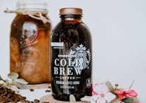 A bottle of Starbucks Cold Brew coffee is prominently displayed alongside a jar filled with coffee beans. Decorative flowers and leaves adorn the scene, adding elegance. Coffee beans are scattered around for an aesthetic touch.