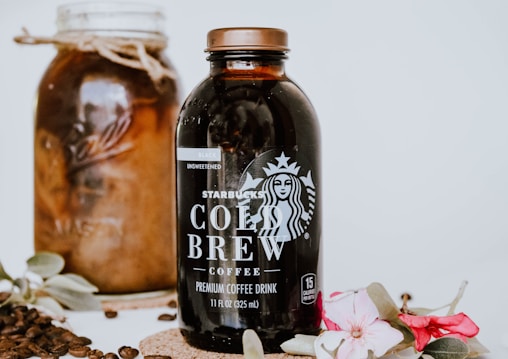 A bottle of Starbucks Cold Brew coffee is prominently displayed alongside a jar filled with coffee beans. Decorative flowers and leaves adorn the scene, adding elegance. Coffee beans are scattered around for an aesthetic touch.