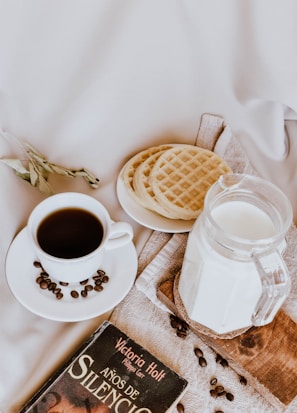 A cozy breakfast setting features a white cup of coffee surrounded by scattered coffee beans, alongside a jug of milk on a wooden board. There are three waffles neatly placed on a white plate, and a book titled 'Años De Silencio' by Victoria Holt is partially visible. The background includes a soft beige fabric, adding to the warm and inviting ambiance.