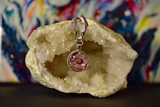 A pendant with a colorful, swirling design is placed in front of a large, open geode with sparkling crystals inside. The pendant is suspended by a purple cord and the background features abstract, vibrant colors in a blurred pattern.