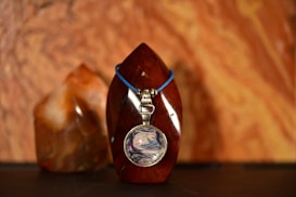 A polished stone sculpture with a smooth surface appears centrally placed, adorned with a pendant on a blue cord. The pendant features a round, colorful design that resembles swirling patterns. Behind it stands another stone of similar texture and color. Both stones are set against a backdrop with earthy tones and fluid patterns.