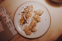 A plate of noodles with various toppings and garnishes, including chopped green onions, nuts, and possibly shredded vegetables or meat. The noodles are artistically arranged on a round, white dish, and the setting includes a menu or notebook with a pencil on the side.