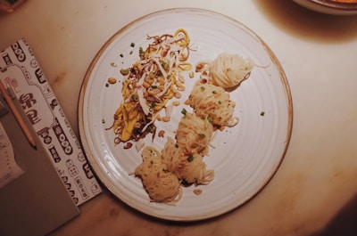 A plate of noodles with various toppings and garnishes, including chopped green onions, nuts, and possibly shredded vegetables or meat. The noodles are artistically arranged on a round, white dish, and the setting includes a menu or notebook with a pencil on the side.