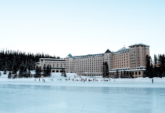 A large, grand hotel with a natural forest backdrop stands prominently. In front of the hotel, a frozen lake serves as an ice skating rink with people enjoying winter activities. The scene is serene with snow surrounding the area and the clear sky enhances the tranquil atmosphere.