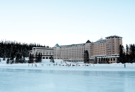 A large, grand hotel with a natural forest backdrop stands prominently. In front of the hotel, a frozen lake serves as an ice skating rink with people enjoying winter activities. The scene is serene with snow surrounding the area and the clear sky enhances the tranquil atmosphere.