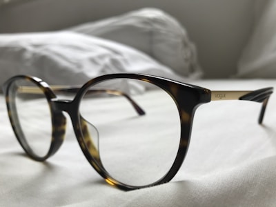 A pair of eyeglasses rests on a light-colored surface, with a focus on the frame and lens details. The frame is made of a tortoiseshell pattern with dark brown and amber tones. The word 'Vogue' is visible on the gold-colored temple arm. The background appears to be soft fabric, suggesting a relaxed or comfortable setting.
