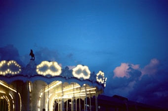 A carousel is illuminated with bright lights against a backdrop of a vibrant evening sky with deep blue and soft pink clouds.