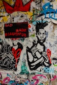 A vibrant collage featuring a mixture of graffiti, poster art, and street art. A prominent artwork of a person wearing boxing gloves labeled with 'Everlast' is central. The phrase 'BAD MADE BEAUTIFUL' appears in bold red and pink letters on a black background. Scattered graffiti and various symbols, including a red heart and a large red crown, cover the walls, all set against a multicolored, textured backdrop.