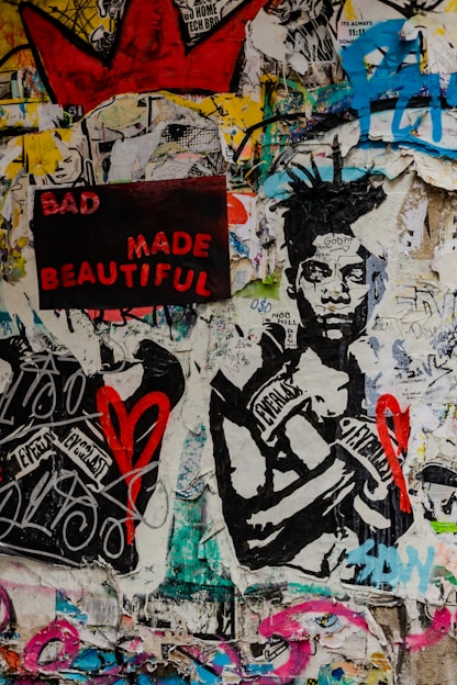 A vibrant collage featuring a mixture of graffiti, poster art, and street art. A prominent artwork of a person wearing boxing gloves labeled with 'Everlast' is central. The phrase 'BAD MADE BEAUTIFUL' appears in bold red and pink letters on a black background. Scattered graffiti and various symbols, including a red heart and a large red crown, cover the walls, all set against a multicolored, textured backdrop.