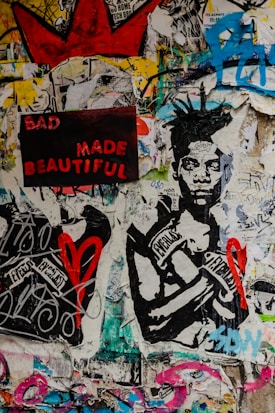 A vibrant collage featuring a mixture of graffiti, poster art, and street art. A prominent artwork of a person wearing boxing gloves labeled with 'Everlast' is central. The phrase 'BAD MADE BEAUTIFUL' appears in bold red and pink letters on a black background. Scattered graffiti and various symbols, including a red heart and a large red crown, cover the walls, all set against a multicolored, textured backdrop.