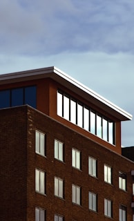 A brick building with a modern penthouse-like extension on the top floor, featuring large windows that reflect the sky. The structure's facade is composed of regular brickwork with symmetrical window placements. The sky is partially overcast, providing a muted backdrop.