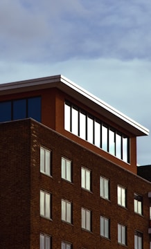 A brick building with a modern penthouse-like extension on the top floor, featuring large windows that reflect the sky. The structure's facade is composed of regular brickwork with symmetrical window placements. The sky is partially overcast, providing a muted backdrop.
