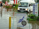 A flooded street with a white van partially submerged in the water and a sign for a diving school in the foreground. There are red umbrellas and outdoor furniture partly submerged, along with several people walking through the water. Signs in multiple languages are visible, indicating a hotel and pharmacy nearby.