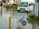 A flooded street with a white van partially submerged in the water and a sign for a diving school in the foreground. There are red umbrellas and outdoor furniture partly submerged, along with several people walking through the water. Signs in multiple languages are visible, indicating a hotel and pharmacy nearby.