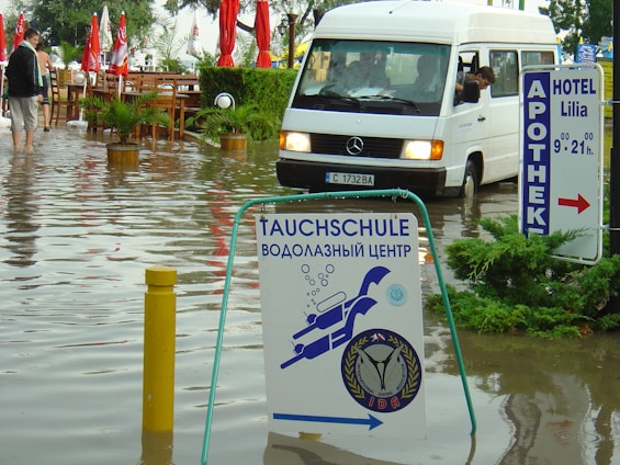 A flooded street with a white van partially submerged in the water and a sign for a diving school in the foreground. There are red umbrellas and outdoor furniture partly submerged, along with several people walking through the water. Signs in multiple languages are visible, indicating a hotel and pharmacy nearby.