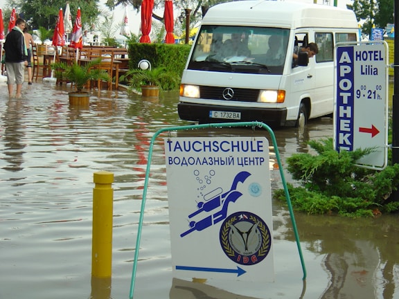 A flooded street with a white van partially submerged in the water and a sign for a diving school in the foreground. There are red umbrellas and outdoor furniture partly submerged, along with several people walking through the water. Signs in multiple languages are visible, indicating a hotel and pharmacy nearby.