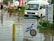 A flooded street with a white van partially submerged in the water and a sign for a diving school in the foreground. There are red umbrellas and outdoor furniture partly submerged, along with several people walking through the water. Signs in multiple languages are visible, indicating a hotel and pharmacy nearby.