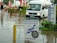 A flooded street with a white van partially submerged in the water and a sign for a diving school in the foreground. There are red umbrellas and outdoor furniture partly submerged, along with several people walking through the water. Signs in multiple languages are visible, indicating a hotel and pharmacy nearby.