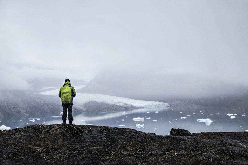 man in green jacket standing on rock formation near body of water