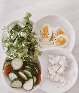 A divided white plate contains four types of food items. In the top left section, there are fresh green lettuce leaves. The top right section holds slices of hard-boiled eggs sprinkled with black pepper. The bottom left section includes sliced cucumbers on a bed of sliced cherry tomatoes. Lastly, the bottom right section has crumbled pieces of feta cheese.