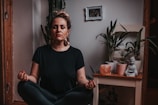 A person is peacefully meditating in a cross-legged position indoors. The surrounding area includes several potted plants, a small Buddha statue, and a salt lamp on a wooden table. A small framed photo is hanging on the wall in the background, adding a personal touch to the serene space.