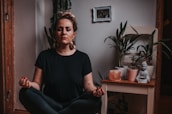 A person is peacefully meditating in a cross-legged position indoors. The surrounding area includes several potted plants, a small Buddha statue, and a salt lamp on a wooden table. A small framed photo is hanging on the wall in the background, adding a personal touch to the serene space.