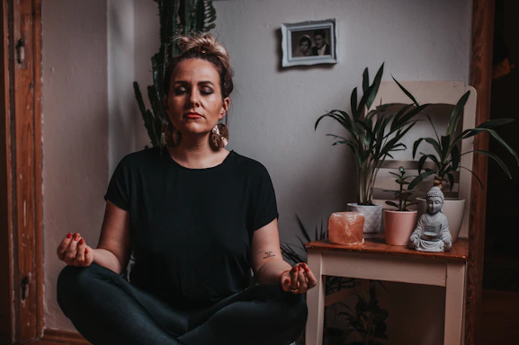 A serene woman meditating in a softly lit room surrounded by plants and calming decor.