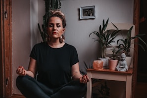 A person is peacefully meditating in a cross-legged position indoors. The surrounding area includes several potted plants, a small Buddha statue, and a salt lamp on a wooden table. A small framed photo is hanging on the wall in the background, adding a personal touch to the serene space.
