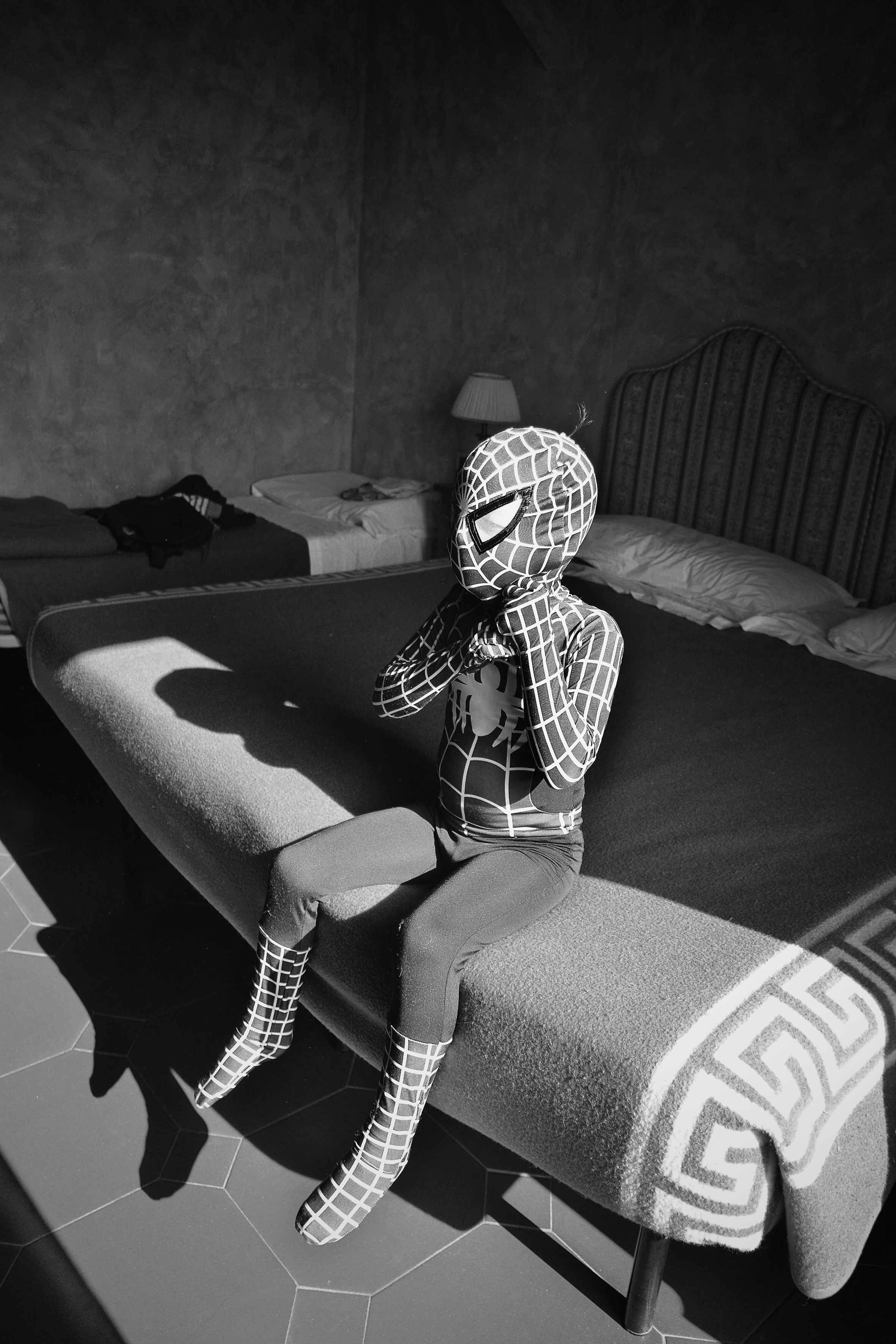 child wearing Spider-Man costume in act of finishing putting on mask while sitting on bed