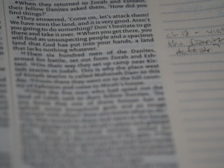 A close-up view of a book page, likely a religious or historical text with printed paragraphs. To the right, there are handwritten notes in blue ink on lined paper.