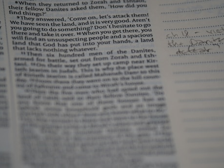A close-up view of a book page, likely a religious or historical text with printed paragraphs. To the right, there are handwritten notes in blue ink on lined paper.