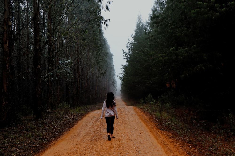 Frau in weißer Jacke geht tagsüber auf Feldweg zwischen grünen Bäumen