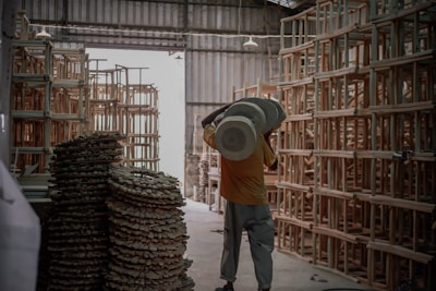 A person is carrying a large cylindrical object in a warehouse filled with stacks of wooden frames and materials. The space is dimly lit, with natural light coming from an open entrance.