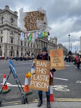 A person stands on a city street holding multiple signs with messages against plastic pollution. The signs are adorned with plastic bottles and bags, emphasizing the environmental protest theme. Surrounding the person are traffic cones and historic buildings under a cloudy sky.