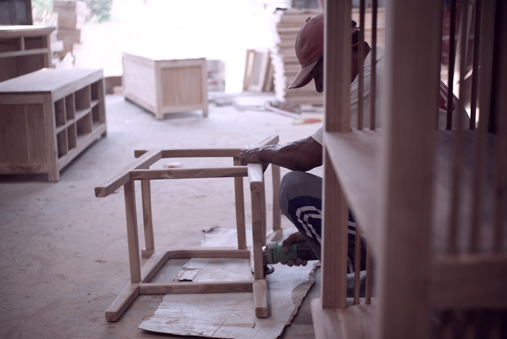 A man in a dusty red baseball cap builds a wooden chair. He is partially hidden by another piece of furniture, but he can be seen holding the frame of the chair in front of him.