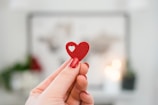 A hand with manicured nails is holding a small red heart with a cut-out in the shape of a smaller heart against a softly blurred background.