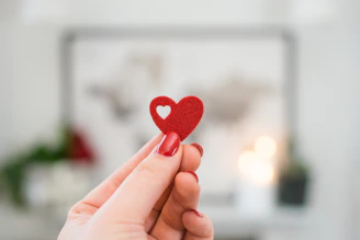 Two hands gently holding a small red heart against a white background.