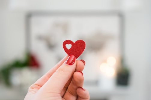 A hand with manicured nails is holding a small red heart with a cut-out in the shape of a smaller heart against a softly blurred background.