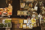 An overhead view of a bustling market stall with various vegetables, fruits, packaged goods, and dried items displayed. There are neatly organized piles of green leafy vegetables, red tomatoes, and potatoes in trays. A lone vendor is seen behind the counter surrounded by scales, bags, and containers. The stall has a traditional, slightly vintage market vibe with labels and signs indicating prices.