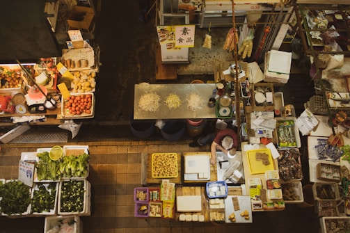 An overhead view of a bustling market stall with various vegetables, fruits, packaged goods, and dried items displayed. There are neatly organized piles of green leafy vegetables, red tomatoes, and potatoes in trays. A lone vendor is seen behind the counter surrounded by scales, bags, and containers. The stall has a traditional, slightly vintage market vibe with labels and signs indicating prices.
