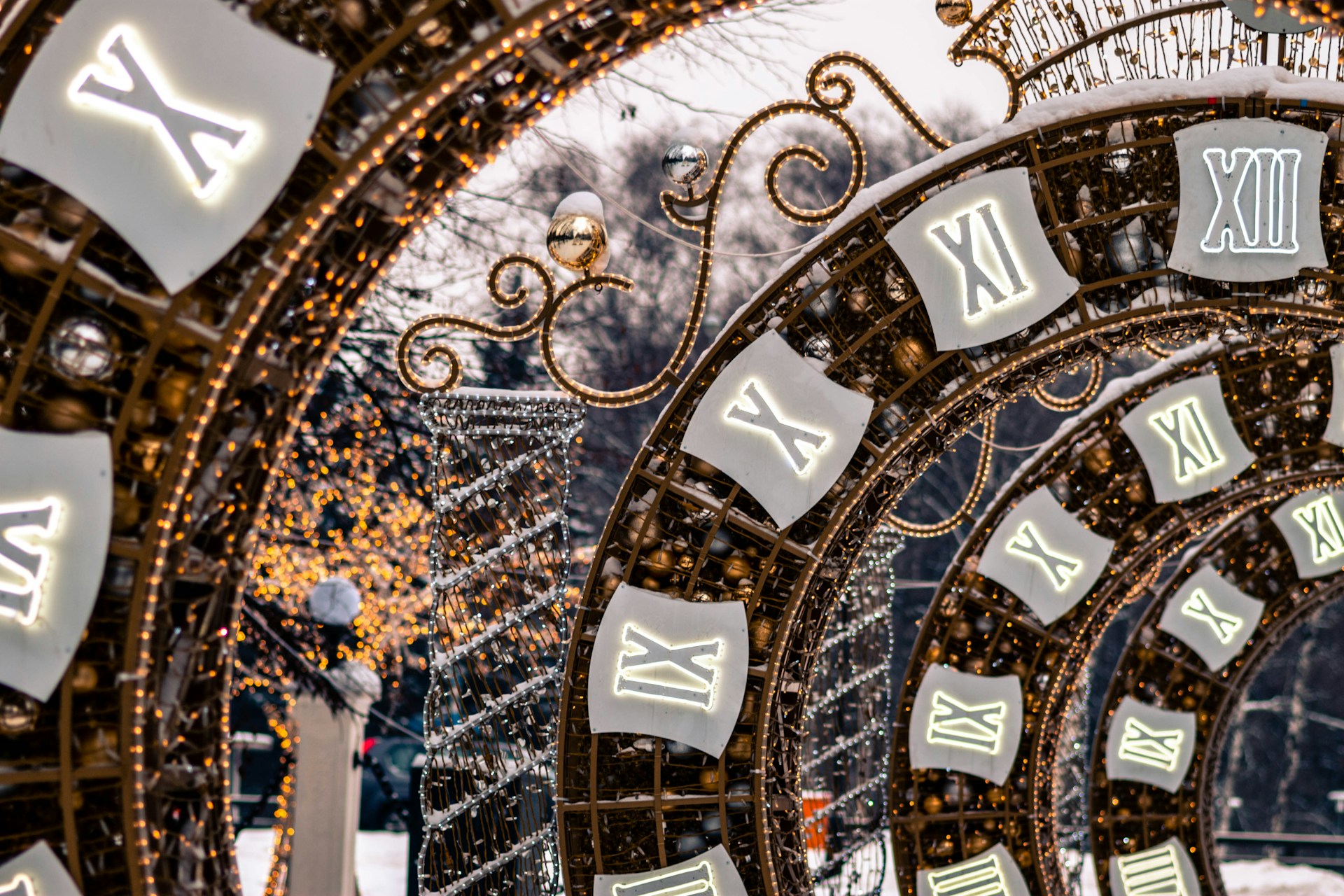 A series of large, decorative arches adorned with illuminated Roman numerals and intricate light fixtures, creating a festive and enchanting atmosphere. The scene is set against a backdrop of trees with twinkling fairy lights, suggesting a holiday or celebratory theme.