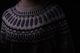 A close-up of a knitted sweater with intricate geometric patterns in black and white. The sweater has a high neckline and showcases various zigzag and dot designs. The lighting is dim, creating a cozy and warm atmosphere.