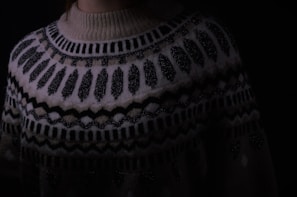 A close-up of a knitted sweater with intricate geometric patterns in black and white. The sweater has a high neckline and showcases various zigzag and dot designs. The lighting is dim, creating a cozy and warm atmosphere.