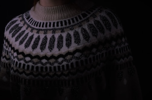A close-up of a knitted sweater with intricate geometric patterns in black and white. The sweater has a high neckline and showcases various zigzag and dot designs. The lighting is dim, creating a cozy and warm atmosphere.