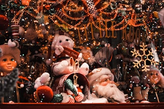 A festive Christmas display filled with toys and decorations. Plushies, nutcrackers, and Santa figurines are surrounded by twinkling lights and colorful ornaments. A sign with 'Little Kook' is featured prominently at the center. Snowflakes and holiday-themed decor create a cheerful and whimsical atmosphere.