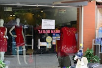 A storefront with glass doors displaying mannequins wearing red dresses. The shop sign reads 'TAILOR MADE' with additional signage about discounts and payment options. A scooter is parked outside along with a shelf of bottled water.
