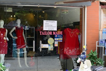 A storefront with glass doors displaying mannequins wearing red dresses. The shop sign reads 'TAILOR MADE' with additional signage about discounts and payment options. A scooter is parked outside along with a shelf of bottled water.