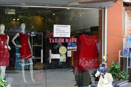 A storefront with glass doors displaying mannequins wearing red dresses. The shop sign reads 'TAILOR MADE' with additional signage about discounts and payment options. A scooter is parked outside along with a shelf of bottled water.